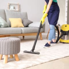 A person wearing yellow gloves and an apron is vacuuming a light-colored rug in a living room with a gray sofa, colorful pillows, and a small knitted stool—a perfect scene for Property Management or Fully Managed Holiday Lets. A yellow vacuum cleaner is nearby.