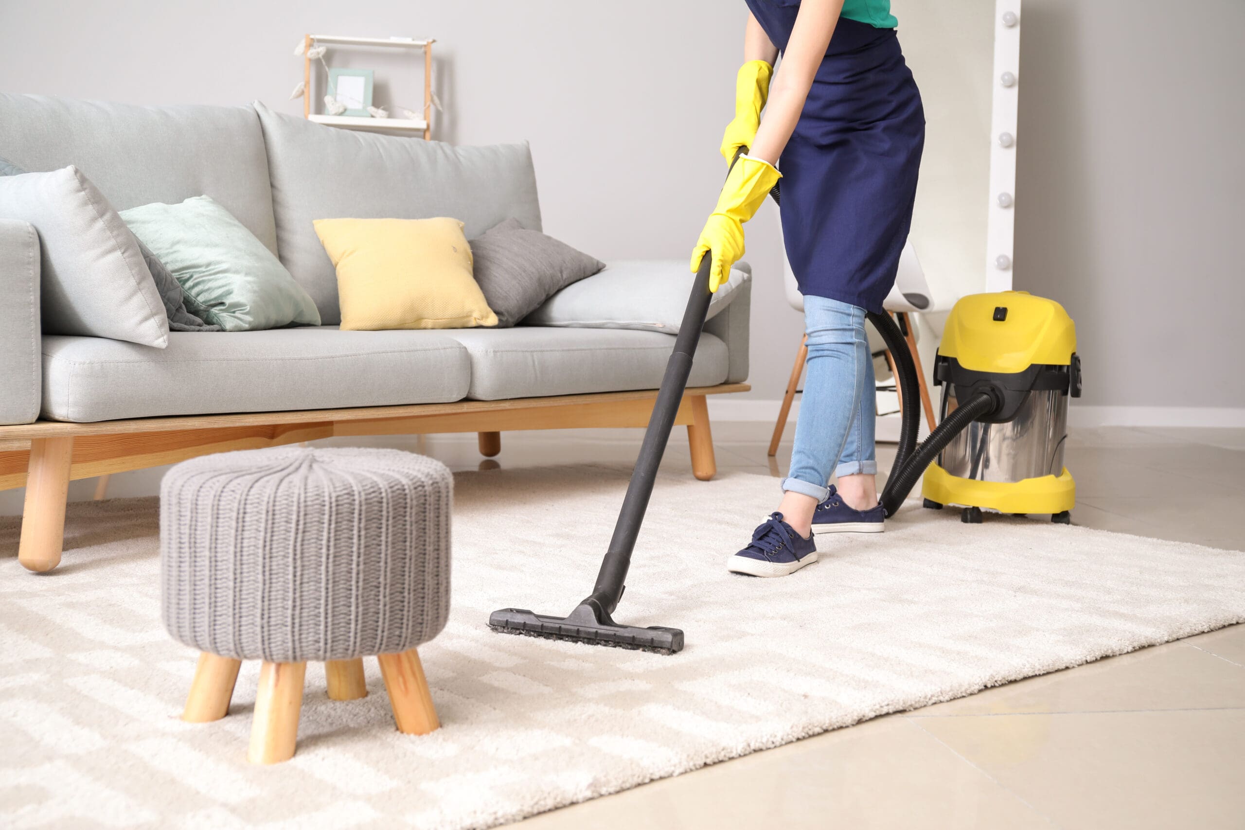 A person wearing yellow gloves and an apron is vacuuming a light-colored rug in a living room with a gray sofa, colorful pillows, and a small knitted stool—a perfect scene for Property Management or Fully Managed Holiday Lets. A yellow vacuum cleaner is nearby.