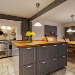 Modern kitchen with gray cabinets, wooden countertops, and stainless steel appliances—perfect for fully managed holiday lets. A vase of yellow flowers sits on the island under pendant lights, with a dining table and fruit in the background.