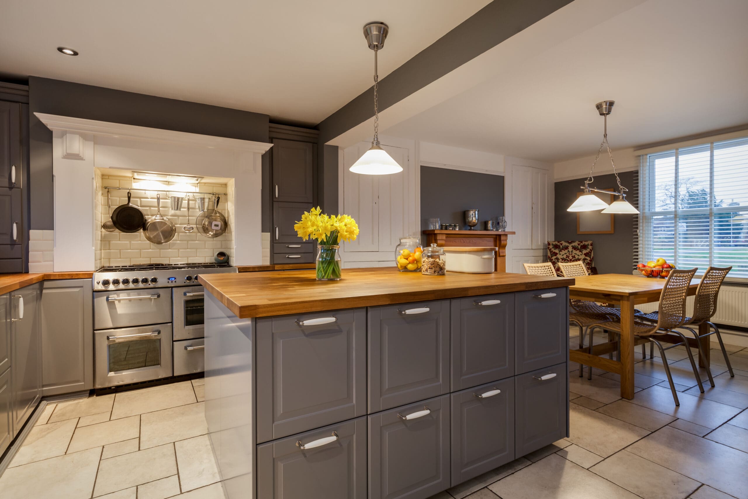 Modern kitchen with gray cabinets, wooden countertops, and stainless steel appliances—perfect for fully managed holiday lets. A vase of yellow flowers sits on the island under pendant lights, with a dining table and fruit in the background.