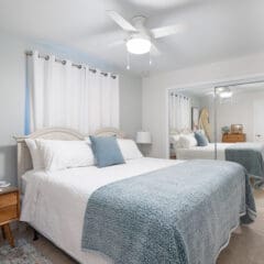 A bright bedroom with a neatly made bed, white and blue bedding, a wooden nightstand with a lamp, ceiling fan, light blue walls, curtains, and a mirrored closet—ideal for Fully Managed Holiday Lets or Second Home Services.