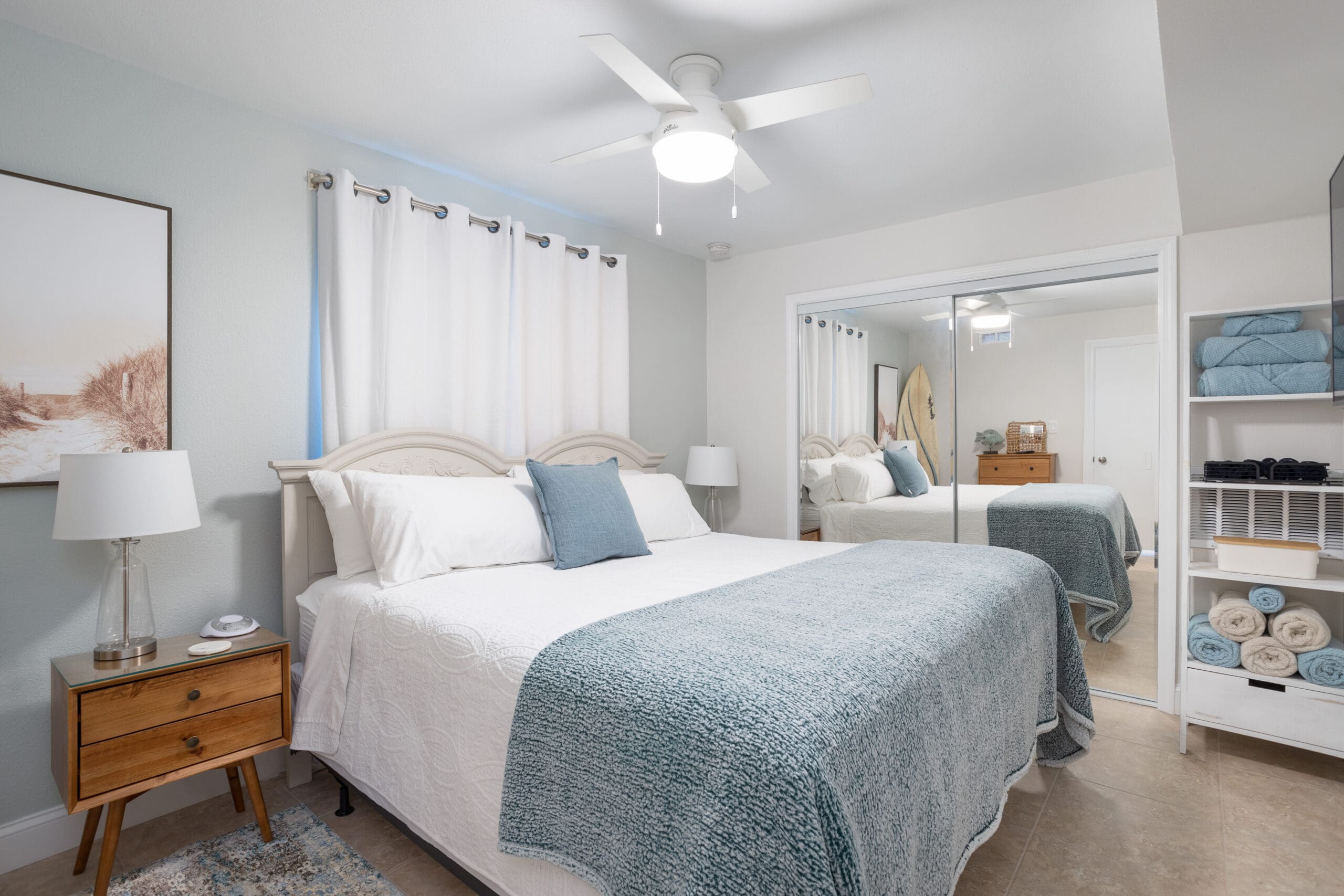 A bright bedroom with a neatly made bed, white and blue bedding, a wooden nightstand with a lamp, ceiling fan, light blue walls, curtains, and a mirrored closet—ideal for Fully Managed Holiday Lets or Second Home Services.