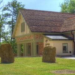 A modern brick house with a steep, tiled roof stands next to a well-kept lawn featuring two large upright stones. Ideal for second home owners seeking Property Management or Holiday Home Cleaning services. Trees and a wooden fence are visible in the background.