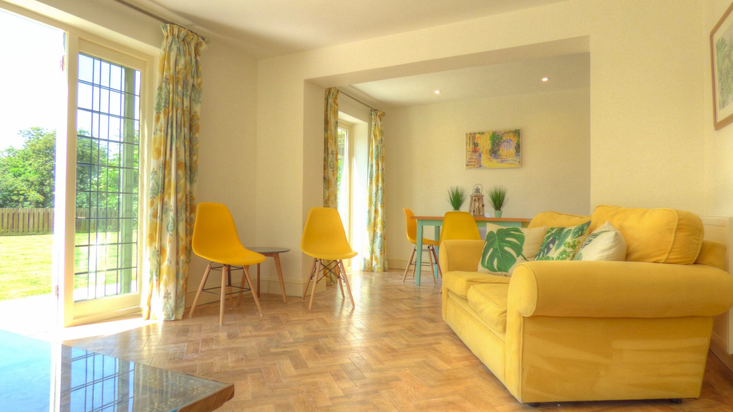 Bright living and dining area with yellow chairs, a yellow sofa with patterned cushions, wooden herringbone floor, large windows, and colorful curtains overlooking a green garden—ideal for effortless property management or second home services.