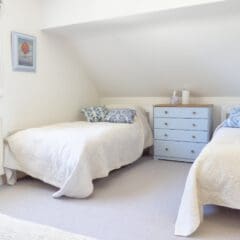 Bright bedroom with three single beds, white bedding, blue and white pillows, a small blue chest of drawers—ideal for Airbnb Management or Second Home Services. Window blinds and a framed picture complete this inviting space.