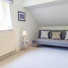 A bright, minimalist room with a gray sofa, three decorative cushions, and a small white side table—perfectly styled for Airbnb Management. A window with striped blinds and a radiator beneath complete this inviting holiday let.