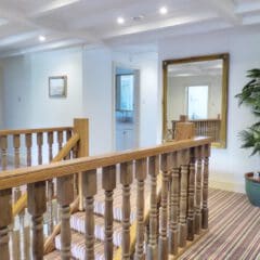A bright hallway with wooden railings, striped carpet, a large gold-framed mirror, framed picture, green potted plant, and a view into a bathroom with white fittings—perfect for Fully Managed Holiday Lets or seamless Airbnb Management.