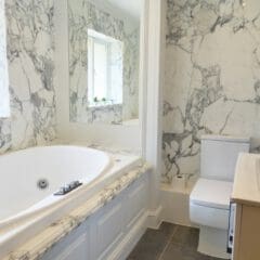 Modern bathroom with white marble walls, built-in bathtub, toilet, rectangular sink, large mirror, and a window lined with potted plants—an inviting space ideal for fully managed holiday lets or seamless Airbnb management. Gray tile flooring completes the look.