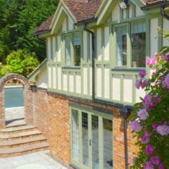 A sunlit brick and timber house with large windows, a garden patio, stone steps, and an arched wall. Pink flowers and green foliage surround the peaceful outdoor space—ideal for fully managed holiday lets.