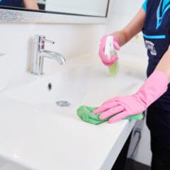 A person wearing pink rubber gloves cleans a bathroom sink with a green cloth and spray bottle—showcasing meticulous Holiday Home Cleaning. The faucet, countertop, and soap container are visible.