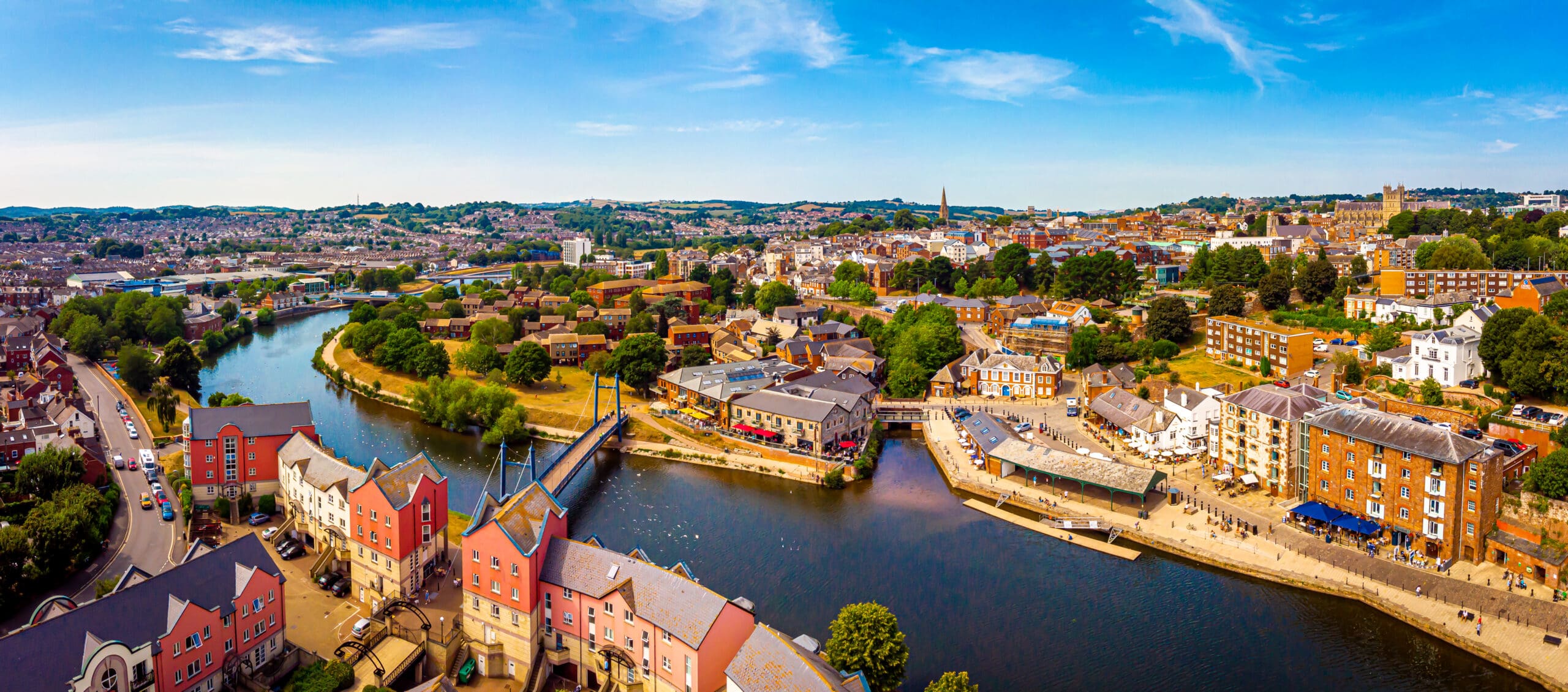 Aerial view of a riverside city with colorful houses, a footbridge, green trees, and clear blue sky on a sunny day—perfect for those seeking fully managed holiday lets or reliable second home services amid stunning scenery.