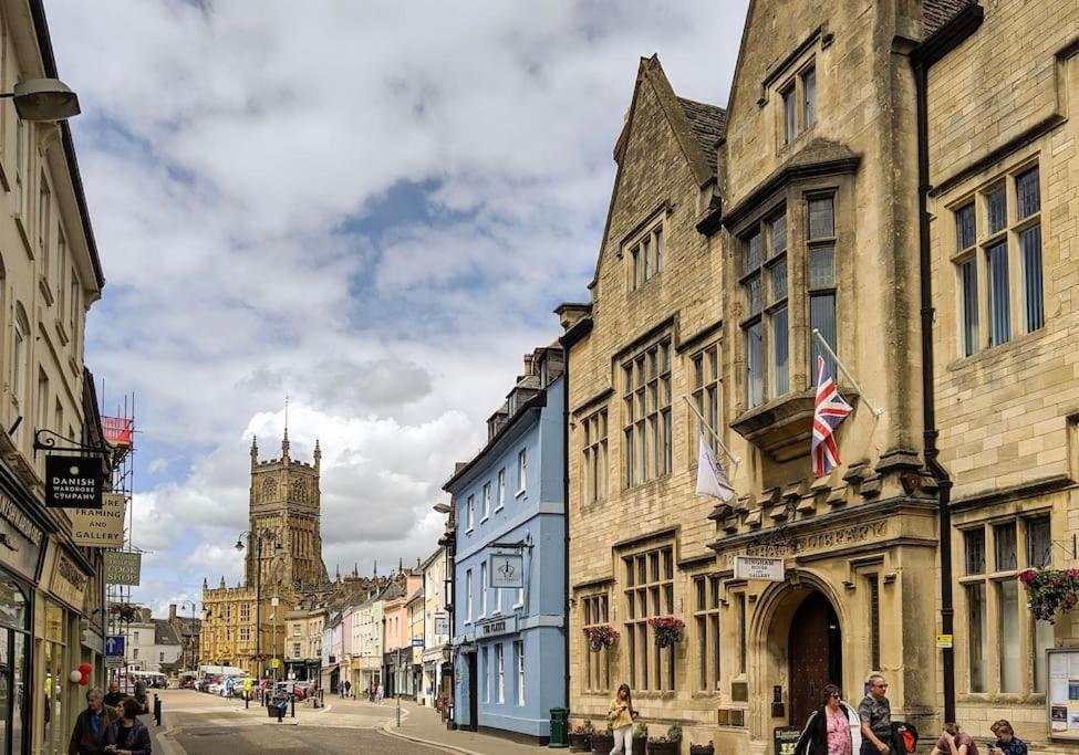 A street scene in an English town with historic stone buildings, a British flag, hanging flower baskets, people walking, and a church tower in the background beneath a partly cloudy sky.