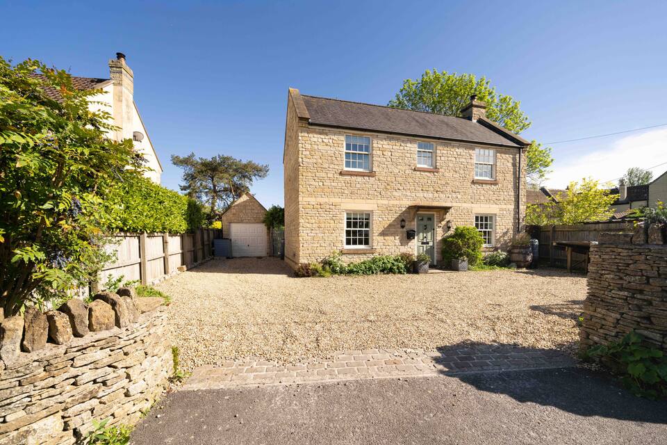 A two-story stone house with a brown roof and white-framed windows stands in a spacious gravel driveway, surrounded by greenery and a low stone wall under a clear blue sky.